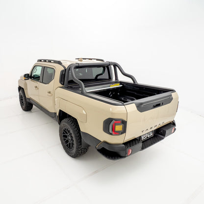 Angled rear view of a Kia Tasman fitted with an HSP Roll R electric roller cover in the open position, showing the aluminium slatted cover retracted at the front of the tub with black sports bars mounted behind the cab.