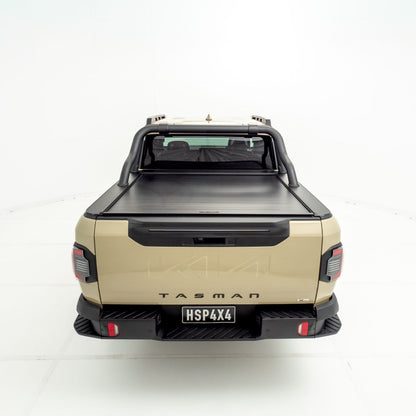 Rear view of a Kia Tasman fitted with an HSP Roll R electric roller cover in the closed position, showing the flat aluminium slatted cover sitting flush across the tub with black sports bars mounted behind the cab.